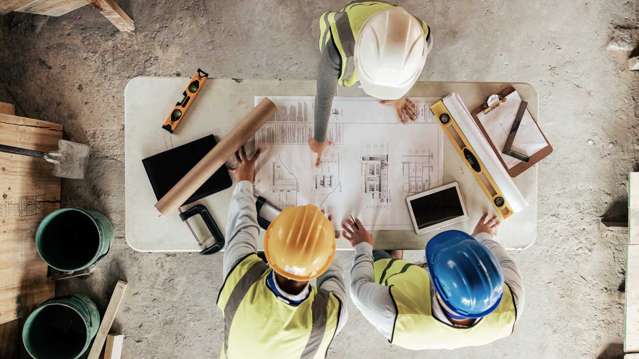 Keeping up With Demand: Three people in safety gear are standing over a table, examining architectural plans at a construction site, surrounded by tools and digital tablets.