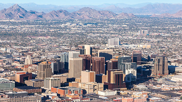Phoenix data center: Aerial view of a dense cityscape with tall buildings, sprawling urban development, distant mountains, and clear skies. No people are visible.