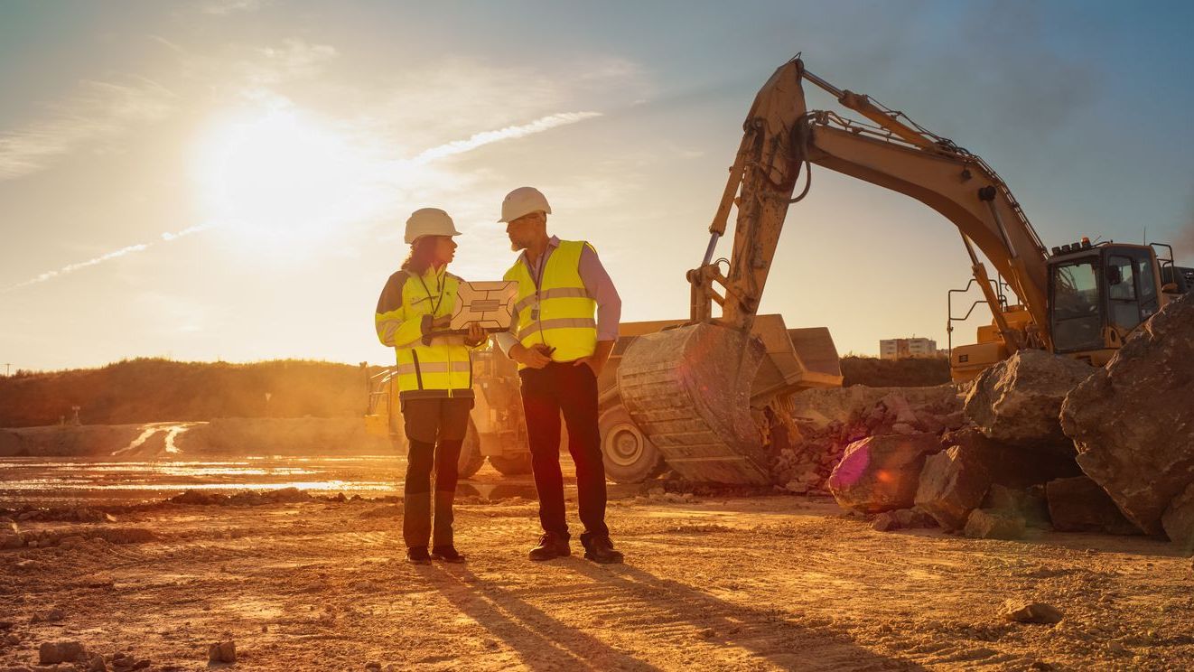 Diversity in construction: Two people wearing high-visibility vests and hard hats discuss plans at a construction site with an excavator and large rocks in the background.