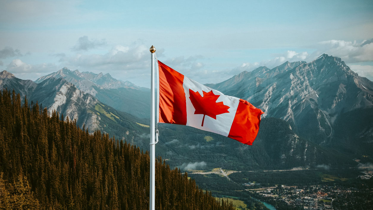 Canadian data center market: A Canadian flag flutters prominently before a stunning mountainous landscape with lush green forests and a small town nestled in the valley below.