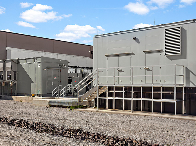 The image shows an industrial building with external ventilation systems, a staircase, and a doorway. It's a sunny day with few clouds in the sky.