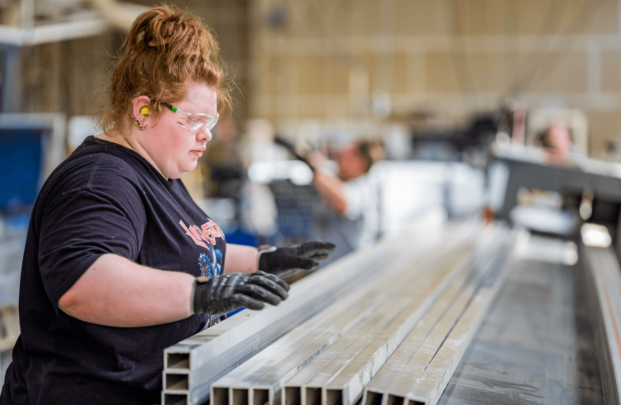 A focused person is inspecting metal profiles while wearing safety glasses, earplugs, and gloves in an industrial setting with coworkers in the background.