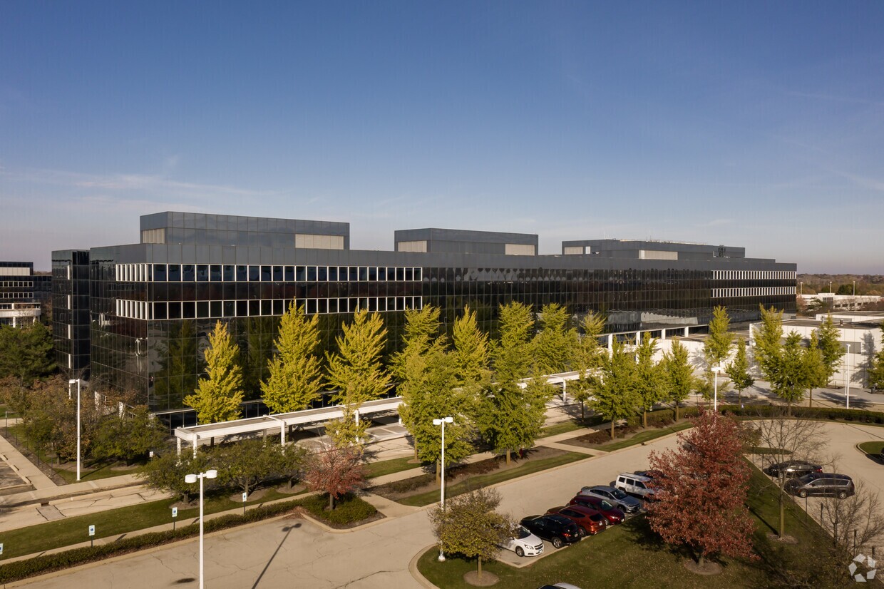 A modern multi-story office building with reflective glass façade surrounded by trees, a parking lot, and a clear sky overhead.