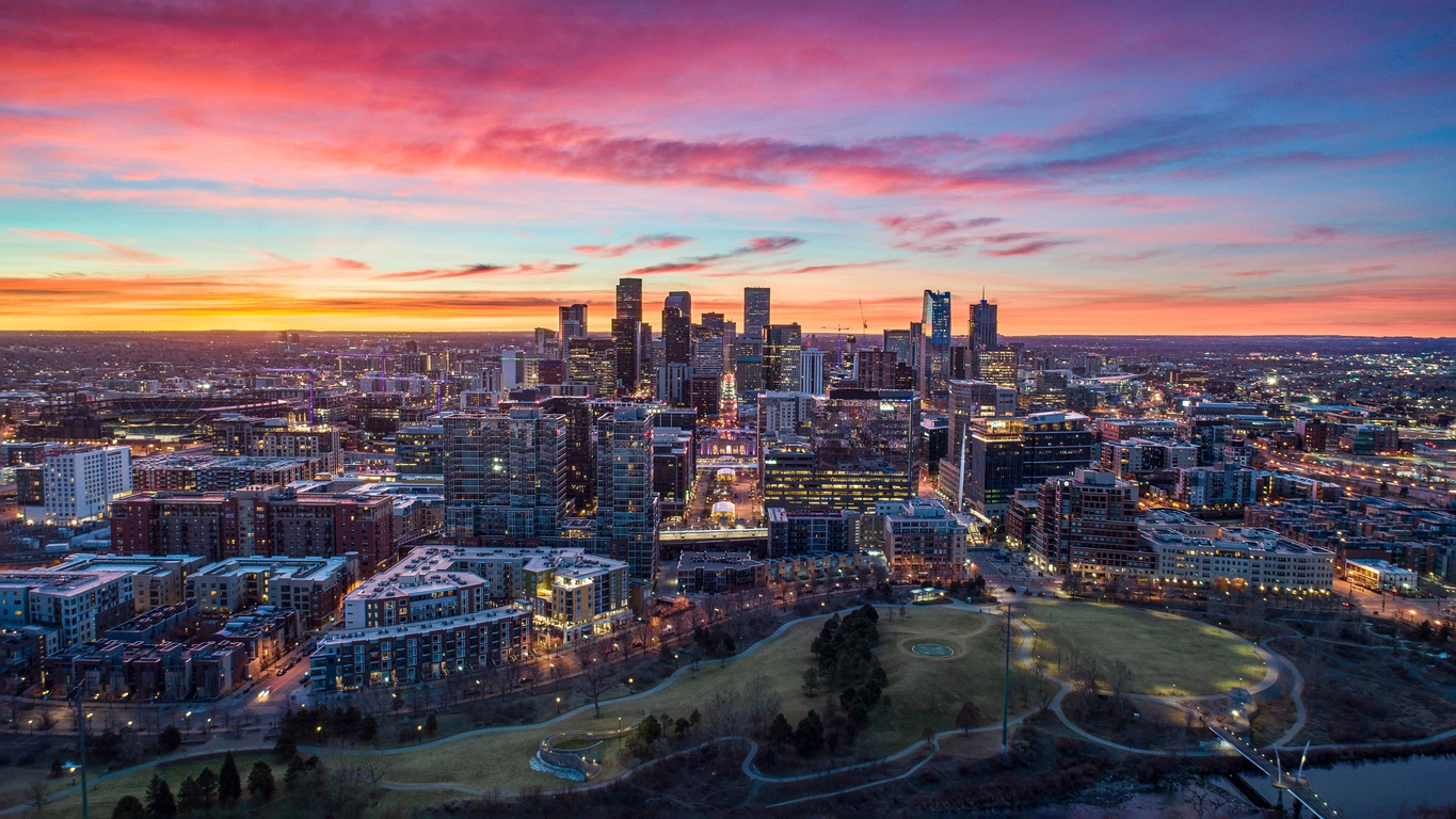 Aerial view of Downtown Denver, Colorado, USA at dusk with vibrant sunset skies, illuminated buildings, a park in the foreground, and sprawling urban development.