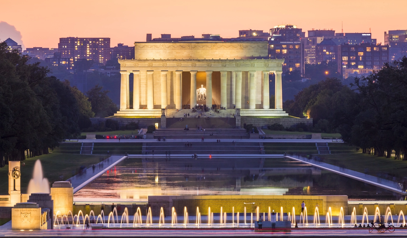 This is an evening shot of the Lincoln Memorial with illuminated fountains in the foreground, reflecting pool, and Washington, DC skyline in the background.