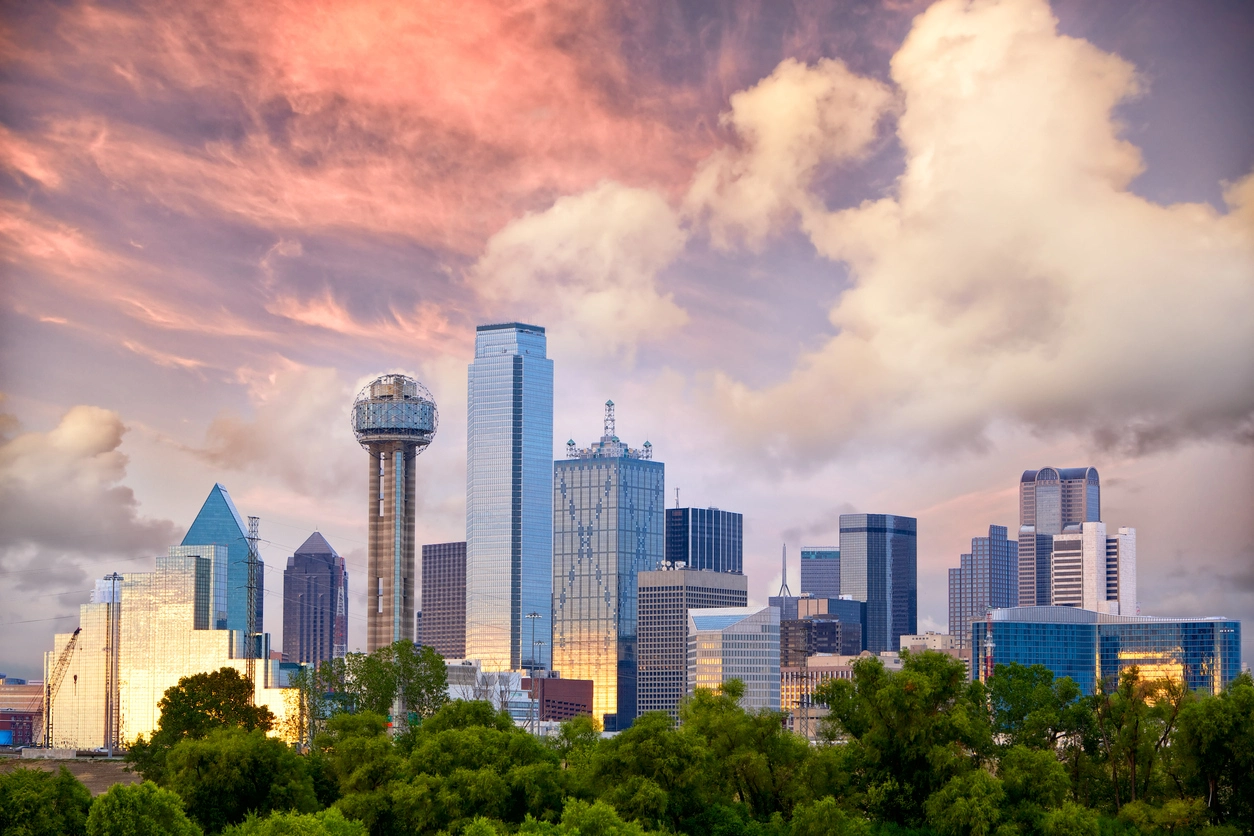 The image shows Dallas skyline against a backdrop of dramatic pink-tinged clouds and blue sky at either sunrise or sunset.