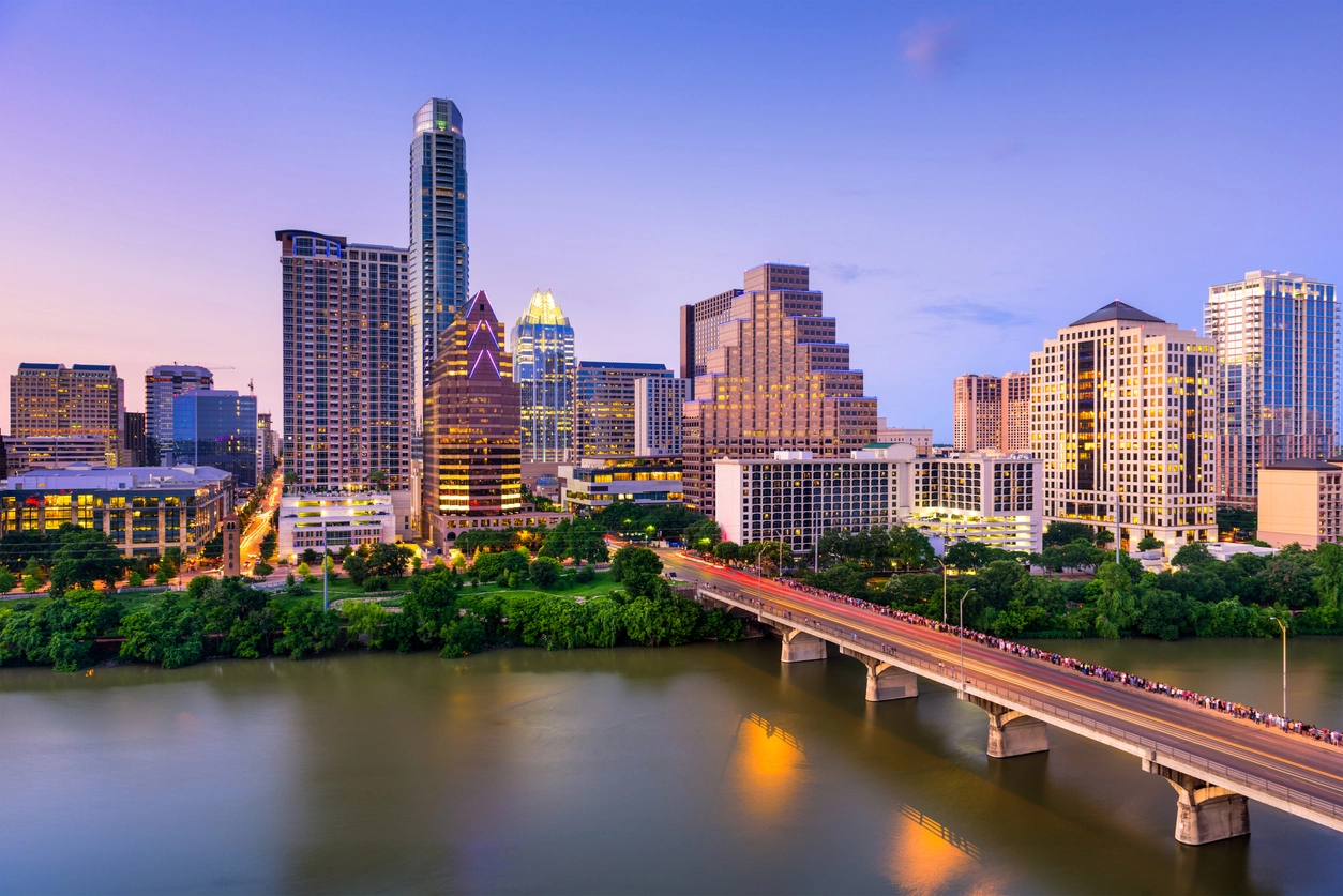 Austin, Texas twilight cityscape with illuminated buildings, bridge over a calm river, clear sky, and dense urban architecture reflected in the water's surface.