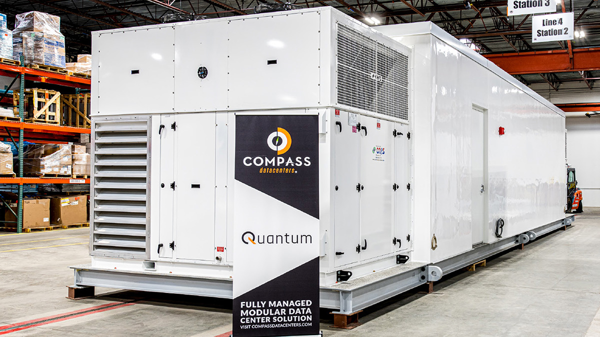 A large, white modular data center unit with 'Compass Quantum' branding sits inside a warehouse, with a forklift visible in the background.