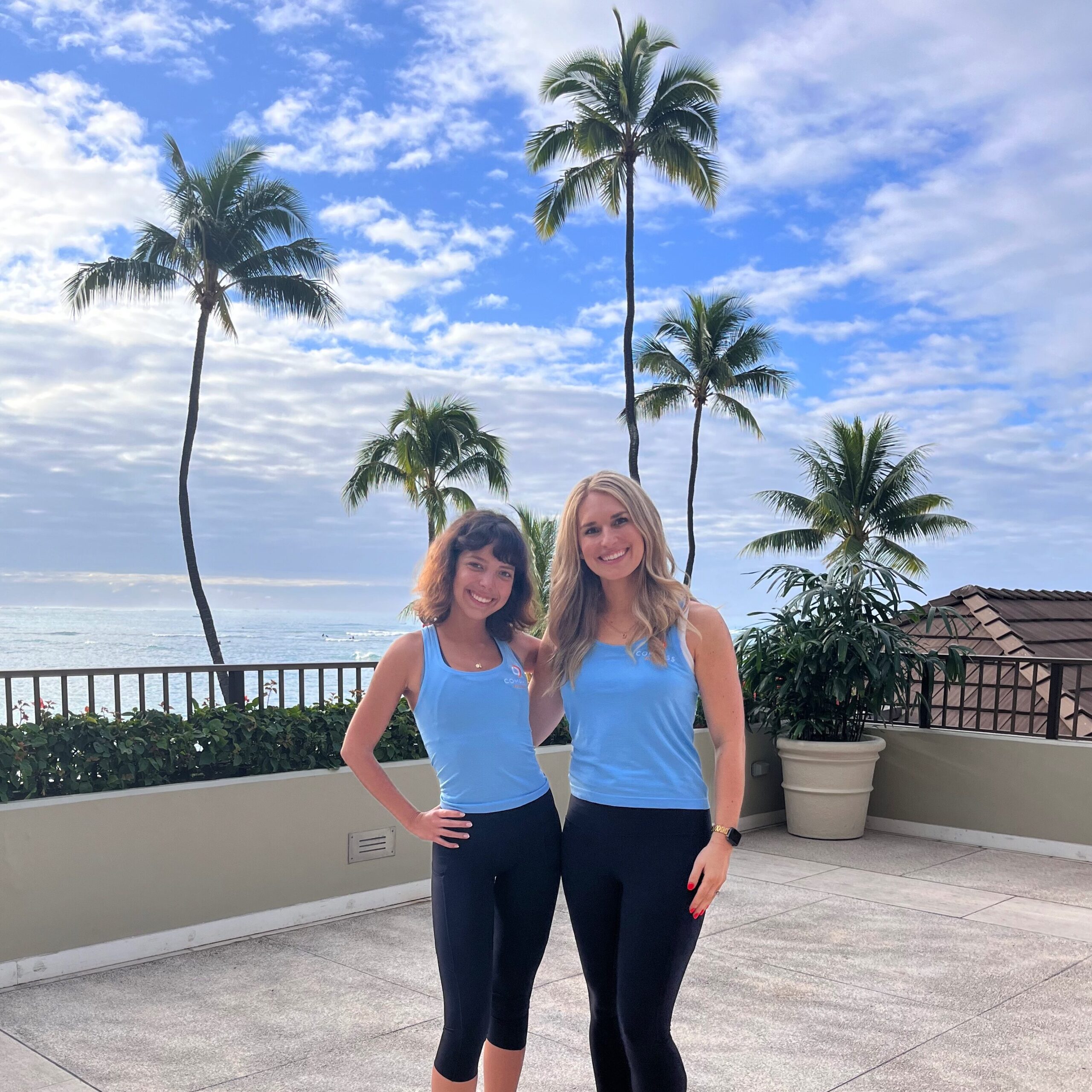 Chelsey Cooper and Frances Verdugo Lash smiling in matching blue tops and black pants on a sunny terrace with palm trees and the ocean in the background.