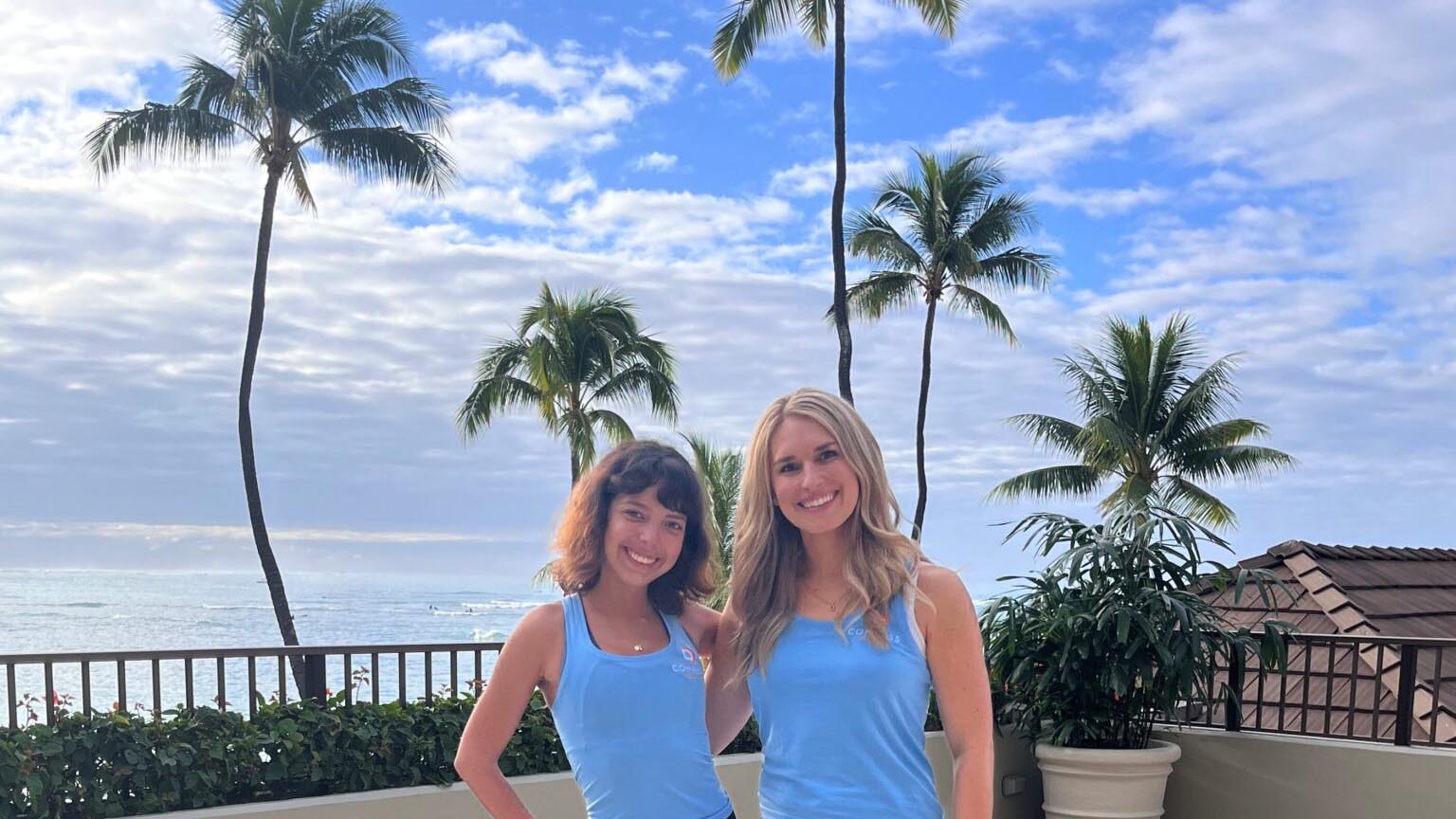 How to support women in the workplace: Two people smiling on a sunny balcony with palm trees, a glimpse of the ocean, and tropical foliage under a partly cloudy sky. They're wearing matching blue tops.