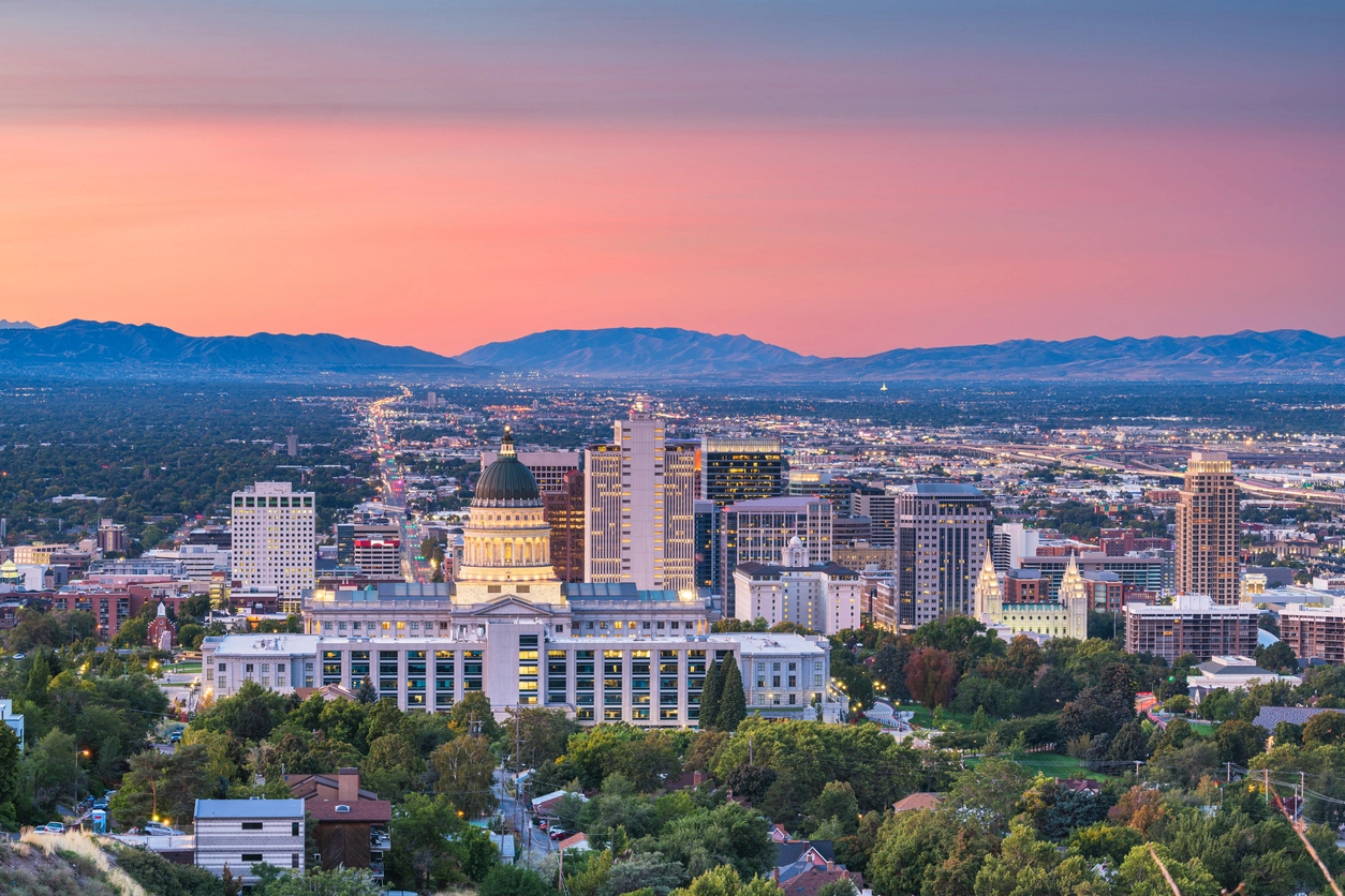 A panoramic view of a city at dusk, featuring a domed capitol building, urban skyline, distant mountains, and a colorful sunset sky.