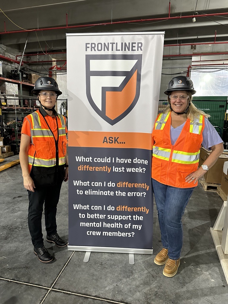 Two people in safety gear stand next to a banner titled "FRONTLINER" with reflective questions focused on improvement and mental health support.